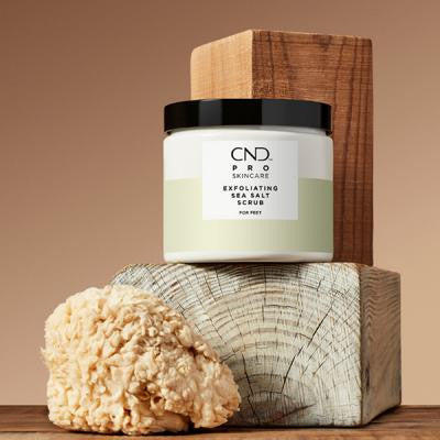 CND Pro Skincare Exfoliating Sea Salt Scrub jar on wood with a natural sponge